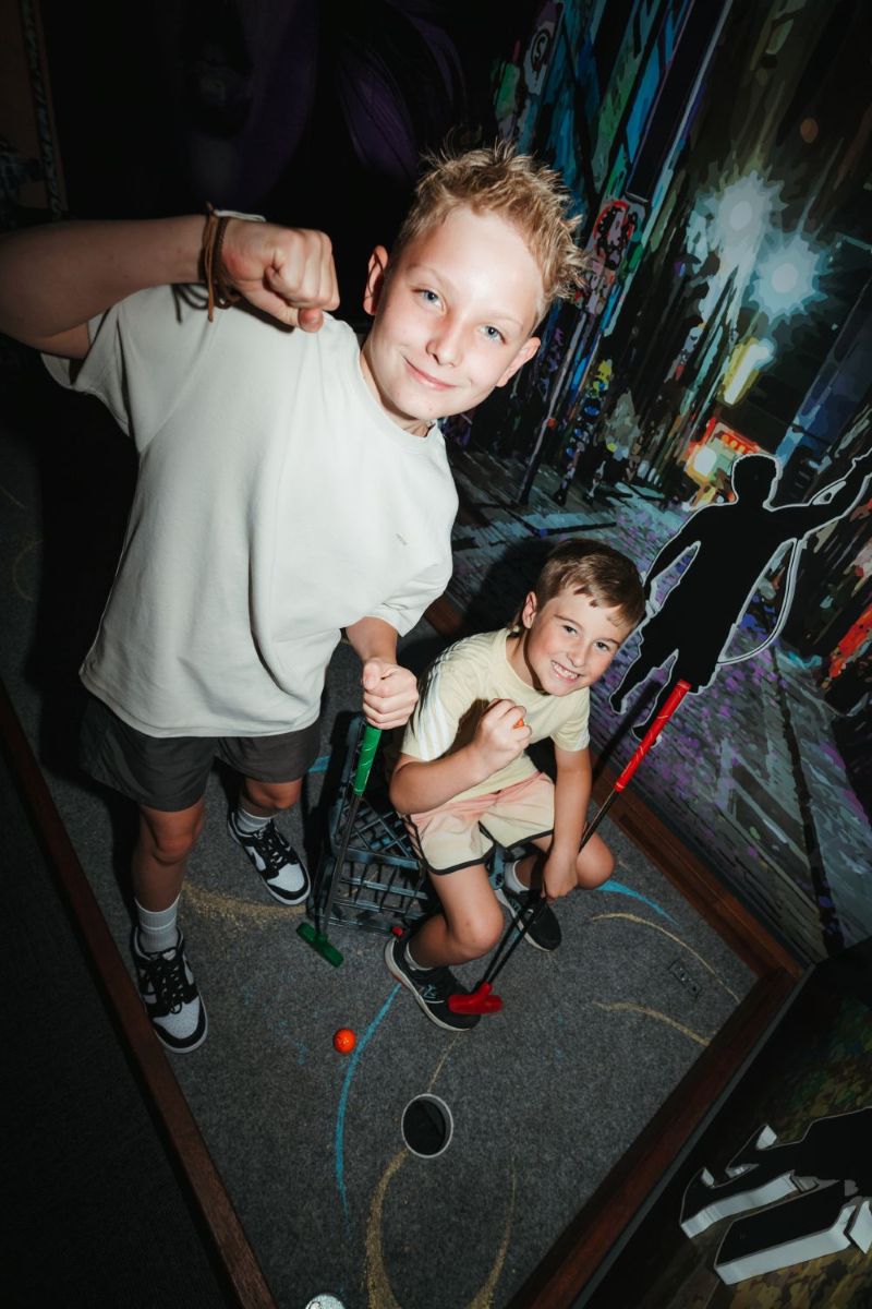 Two boys play together at a colorful miniature golf course, smiling as they take turns putting.