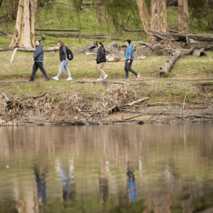 Four people walking along a riverbank, surrounded by trees in the background.