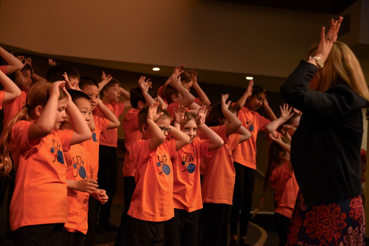 Children in orange shirts and black jackets stand together in front of a stage, smiling and looking excited.