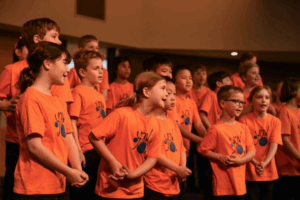 A group of children in orange shirts joyfully singing together in a lively outdoor setting.