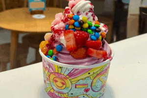 A cup of frozen yoghurt topped with vibrant, colorful sprinkles and a cherry on top, set against a neutral background.