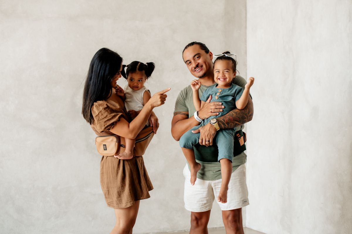 A man and woman stand together, each holding a child in a carrier, smiling in a sunny outdoor setting.
