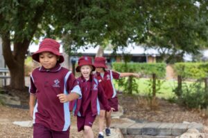 Three children in maroon uniforms walking along a path surrounded by greenery.