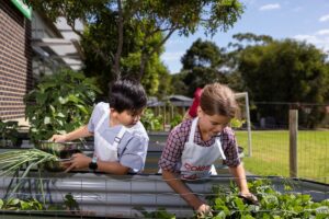 Two children in colorful aprons planting seeds in a garden, surrounded by soil and green plants.