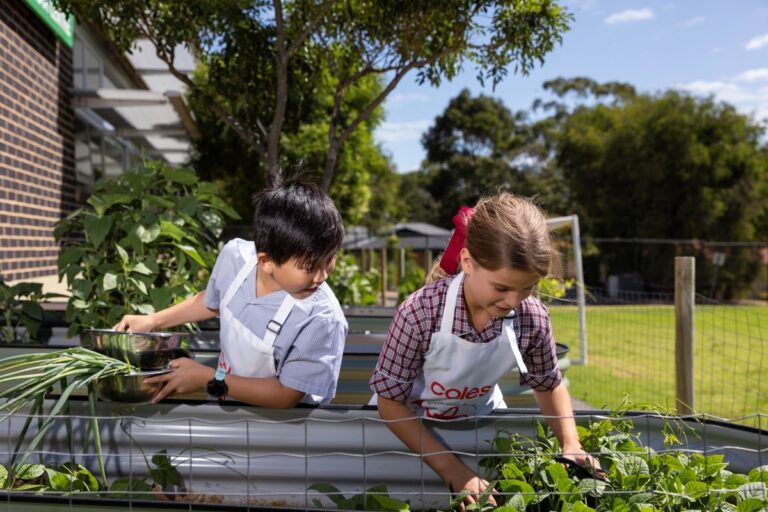 Two children in colorful aprons planting seeds in a garden, surrounded by soil and green plants.