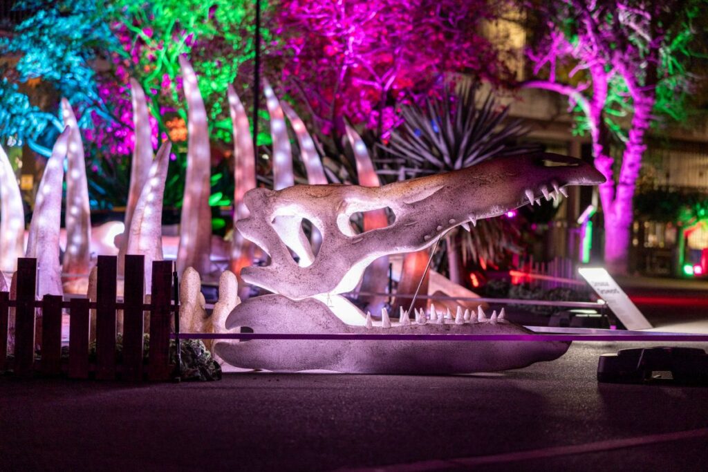 A brightly lit dinosaur statue stands against a dark night sky, showcasing its intricate details and vibrant colors.