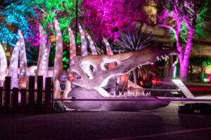 A brightly lit dinosaur statue stands against a dark night sky, showcasing its intricate details and vibrant colors.