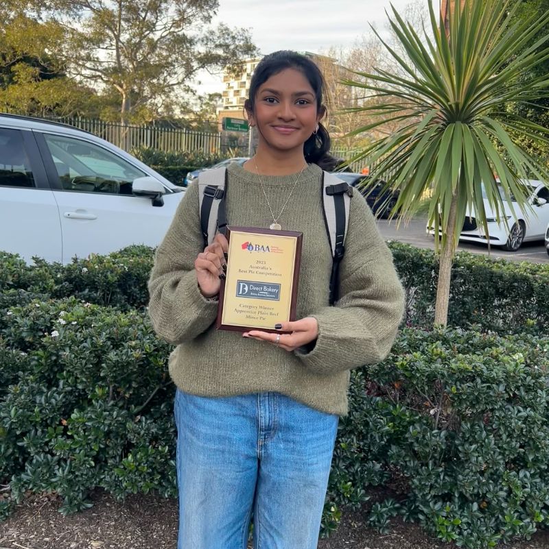 A young woman proudly holds an award plaque, standing in front of lush green bushes.