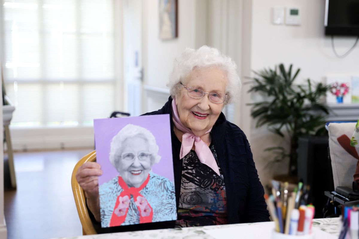 An elderly woman proudly displays a painting of herself, smiling warmly at the viewer.