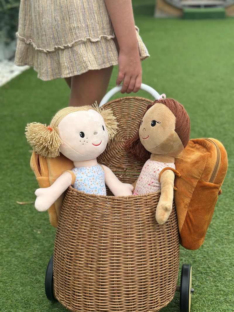 Two dolls nestled in a wicker basket, resting on a green lawn under natural sunlight.
