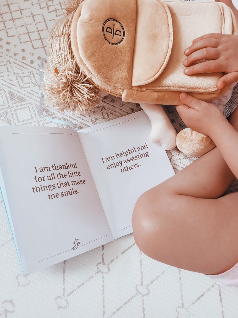 A child sits on the floor, reading a book while holding a stuffed animal close to them.

