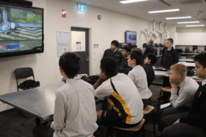 A group of students gathered around a television, intently watching a video game being played.