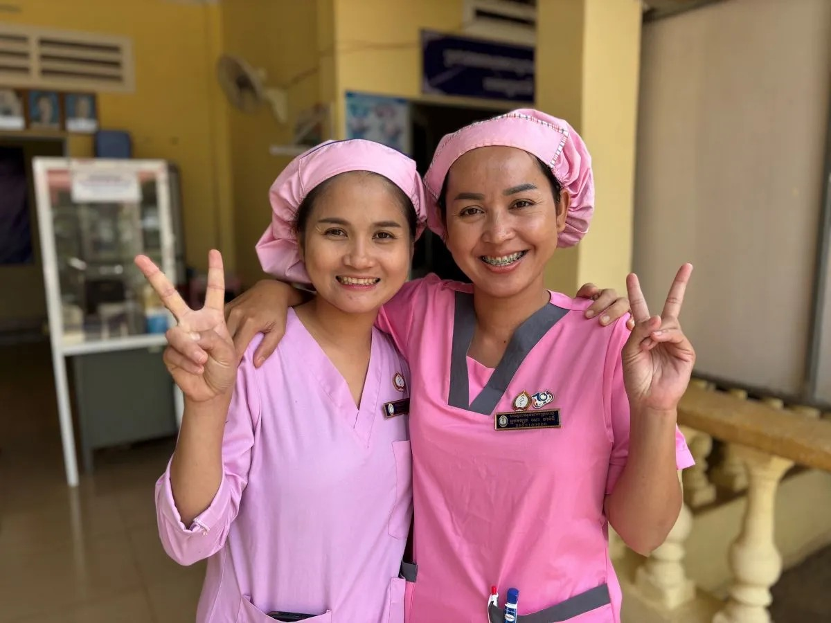 Two women in pink scrubs smile and show the peace sign, conveying a message of positivity and camaraderie.