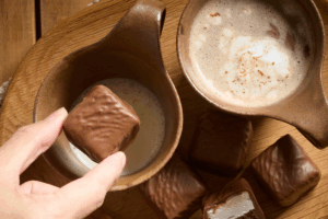 A person holds a chocolate bar in one hand and a cup of coffee in the other, smiling contentedly.