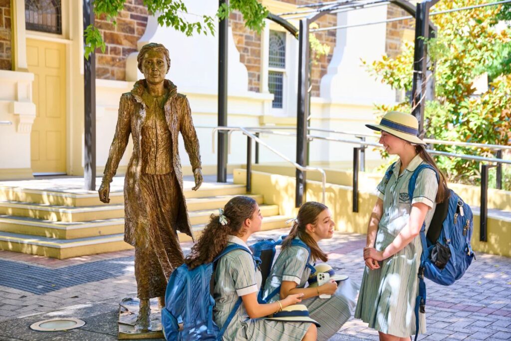 Three girls in school uniforms sit on the ground beside a statue, smiling and chatting in a sunny outdoor setting.