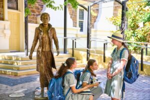 Three girls in school uniforms sit on the ground beside a statue, smiling and chatting in a sunny outdoor setting.