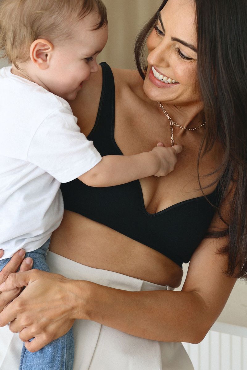 A woman in a black bra top gently holds a baby in her arms, smiling softly at the child.