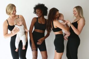 Four women in black leggings and bras, each holding a baby, smiling together in a supportive and joyful setting.