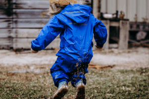 A little girl in a blue rain suit joyfully jumps in a puddle, splashing water around her.