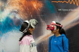 Two girls wearing virtual reality headsets stand in front of a wall, engaged in an immersive digital experience.