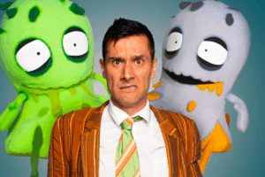 A man in a suit and tie stands beside three colorful stuffed monsters, smiling at the camera.