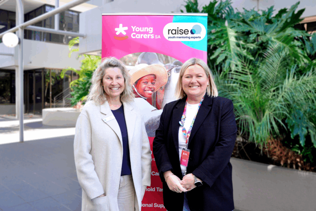 Two women stand beside a pink banner, smiling and engaging with the audience at an event.