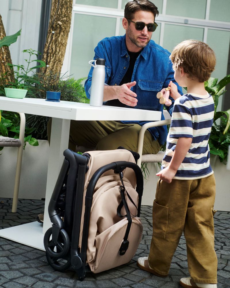 A man and a child stand beside a table with a folded pram, smiling and looking at each other in a bright room.