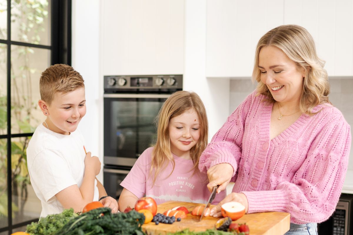 A woman and two children are chopping vegetables together in a bright kitchen, fostering a collaborative cooking experience.