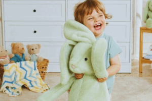 A young child smiles while holding a soft green stuffed rabbit in their arms.