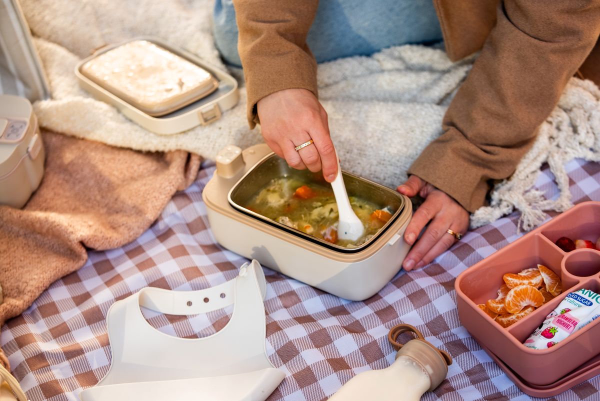 A person enjoys soup from a lunch box while seated on a blanket outdoors.