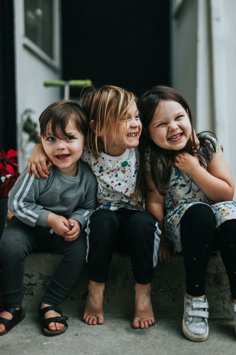 Three young children sit on steps, laughing together and enjoying a joyful moment in the sunlight.