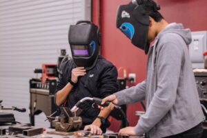 Two men in welding helmets collaborate on a machine, focused on their work in a workshop environment.