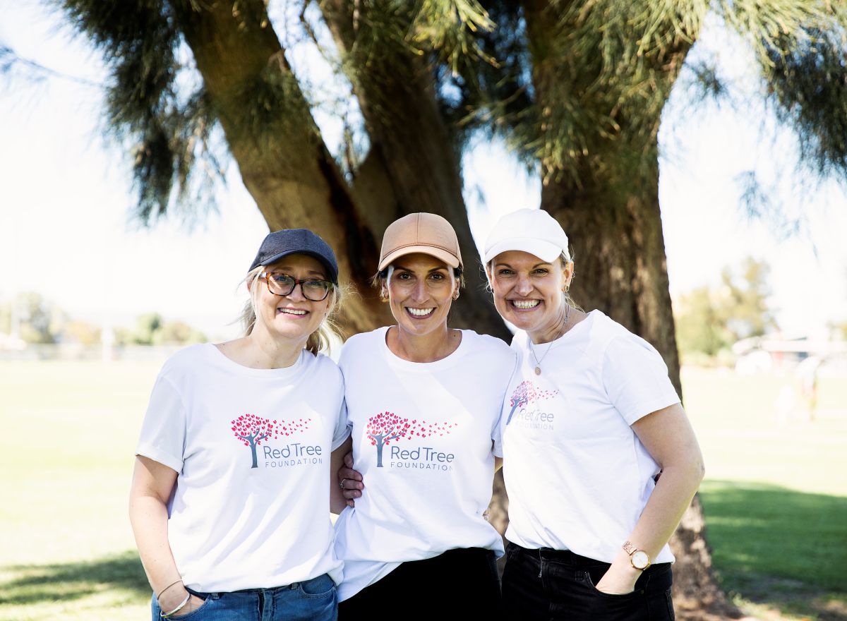 Three women in white shirts and hats stand side by side, smiling at the camera in a bright outdoor setting.
