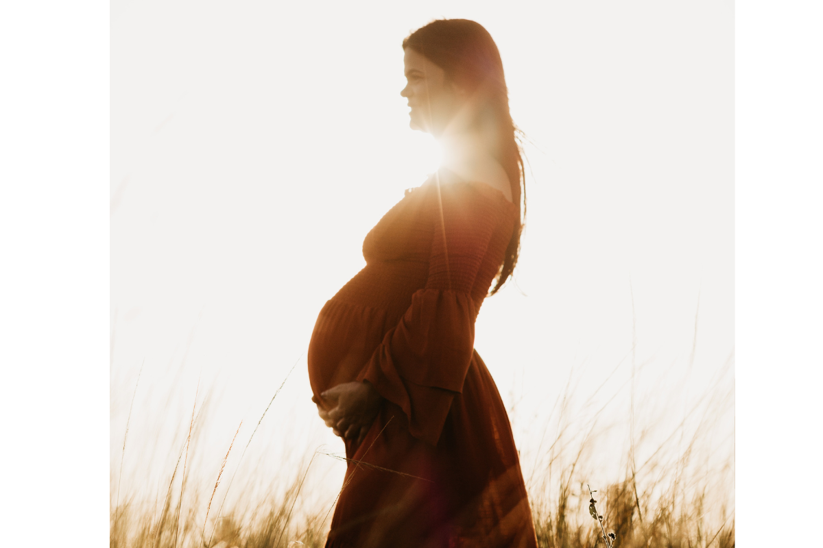 A pregnant woman in a red dress stands gracefully in a lush green field, enjoying the serene outdoor setting.