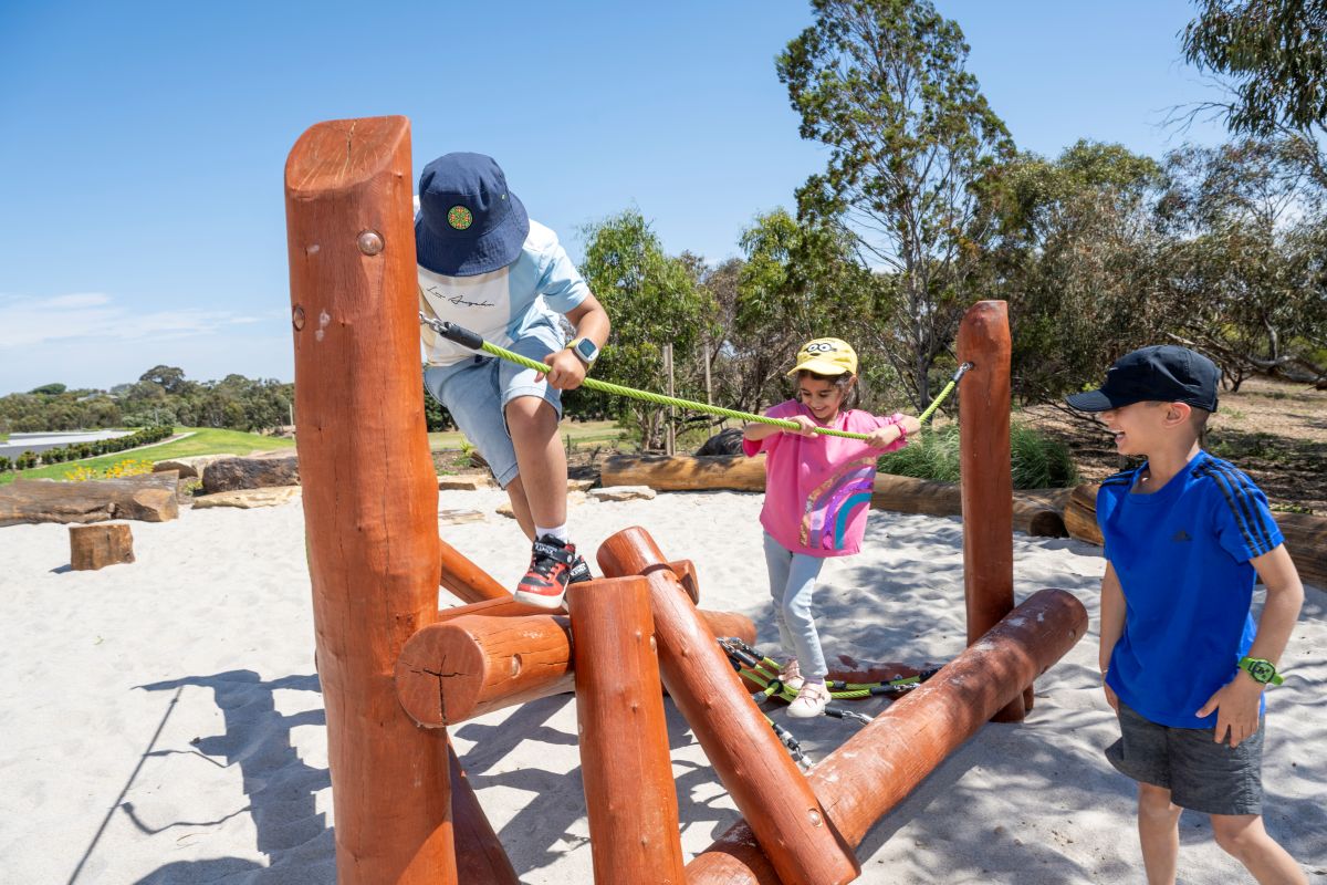 Three children play on a wooden structure, surrounded by sand, enjoying a sunny day.