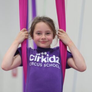 A young girl clutches a purple aerial, highlighting her engagement in an aerial dance or gymnastics activity.