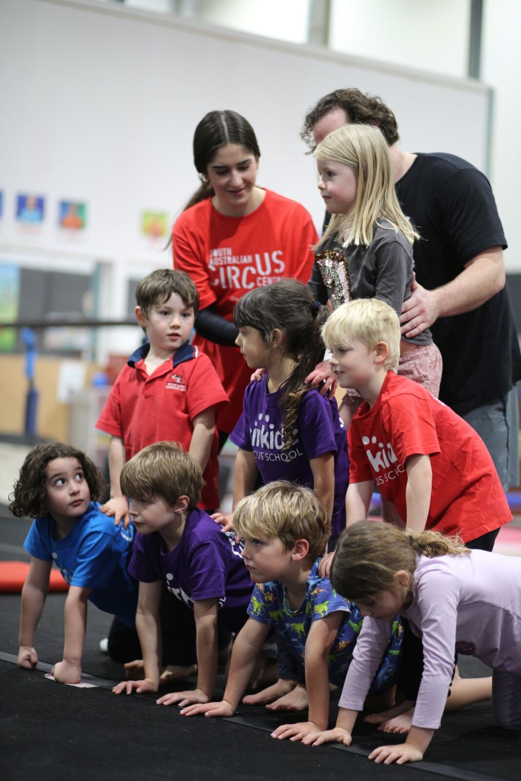 A group of children standing together on a black mat, smiling and engaging with each other.