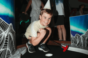 A young boy plays with a golf ball in a colorful game room filled with various toys and games.