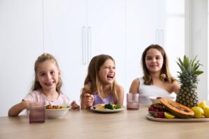 Three girls enjoying a meal at a table filled with fresh fruit and vegetables.