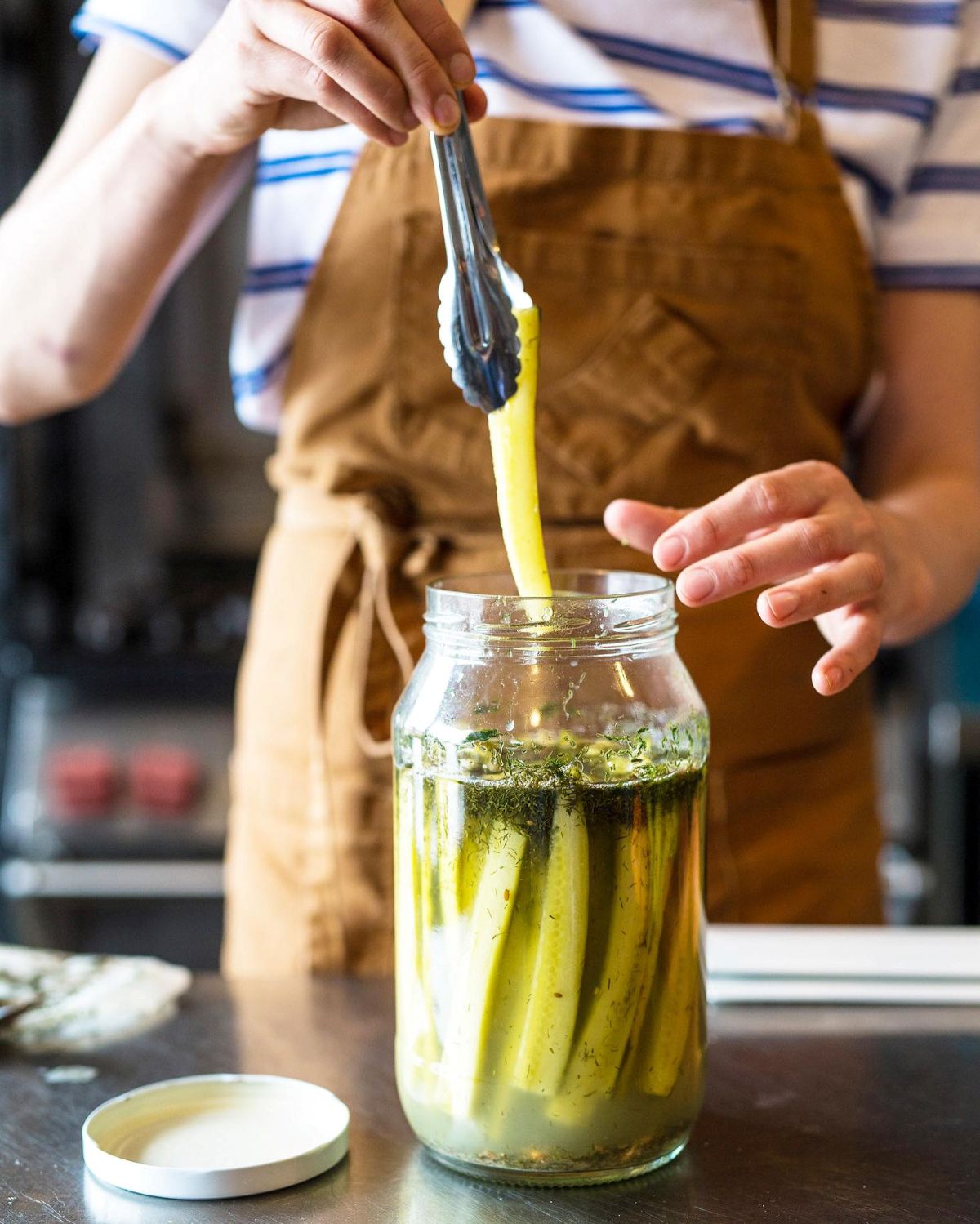 A woman is carefully preparing pickles in a glass jar, surrounded by fresh vegetables and spices.