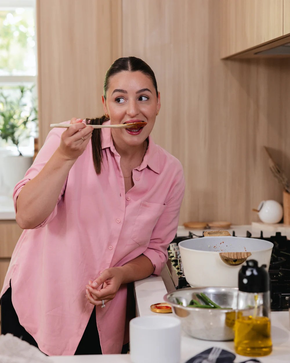 A woman in a pink shirt tastes her food with a wooden spoon.