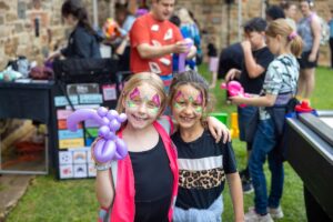 Two young girls with colorful face paint hold bright balloons, smiling joyfully at a festive outdoor event.