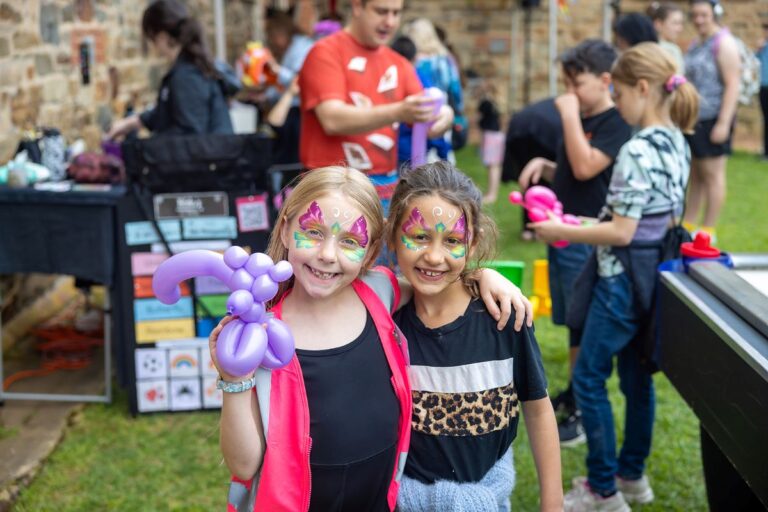 Two young girls with colorful face paint hold bright balloons, smiling joyfully at a festive outdoor event.