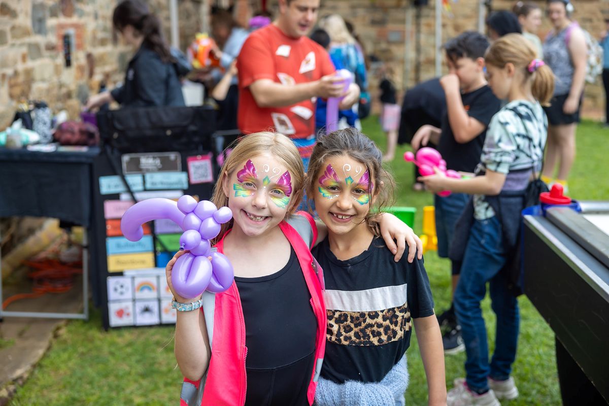 Two young girls with colorful face paint hold bright balloons, smiling joyfully at a festive outdoor event.