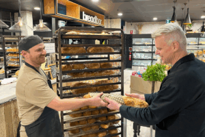 Two men stand in a bakery, one handing a loaf of bread to the other with a friendly expression.