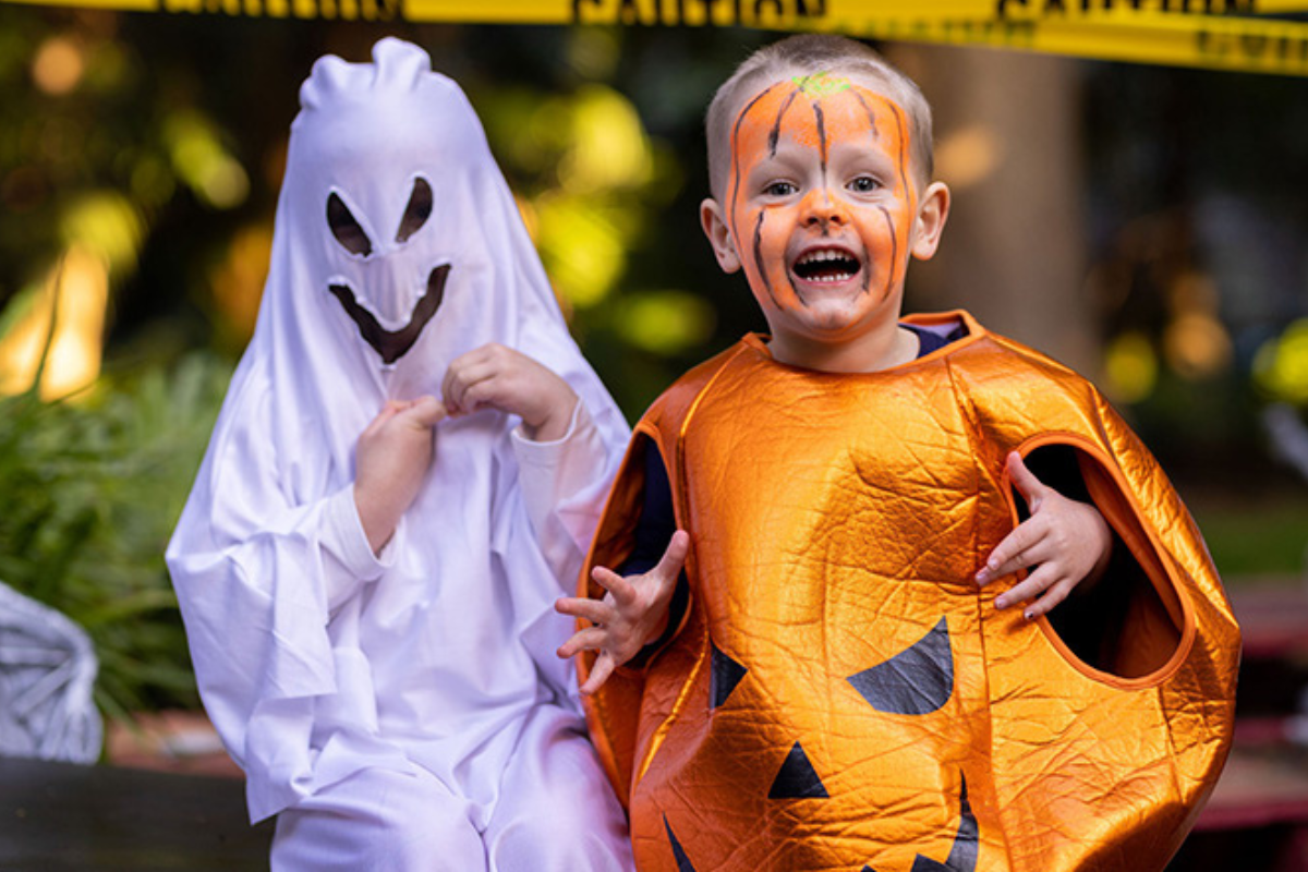 Two children in colorful Halloween costumes, one as a witch and the other as a superhero, smiling and posing together.
