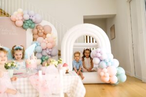 A little girl sits happily under a pink and white balloon arch, smiling and enjoying a festive atmosphere.