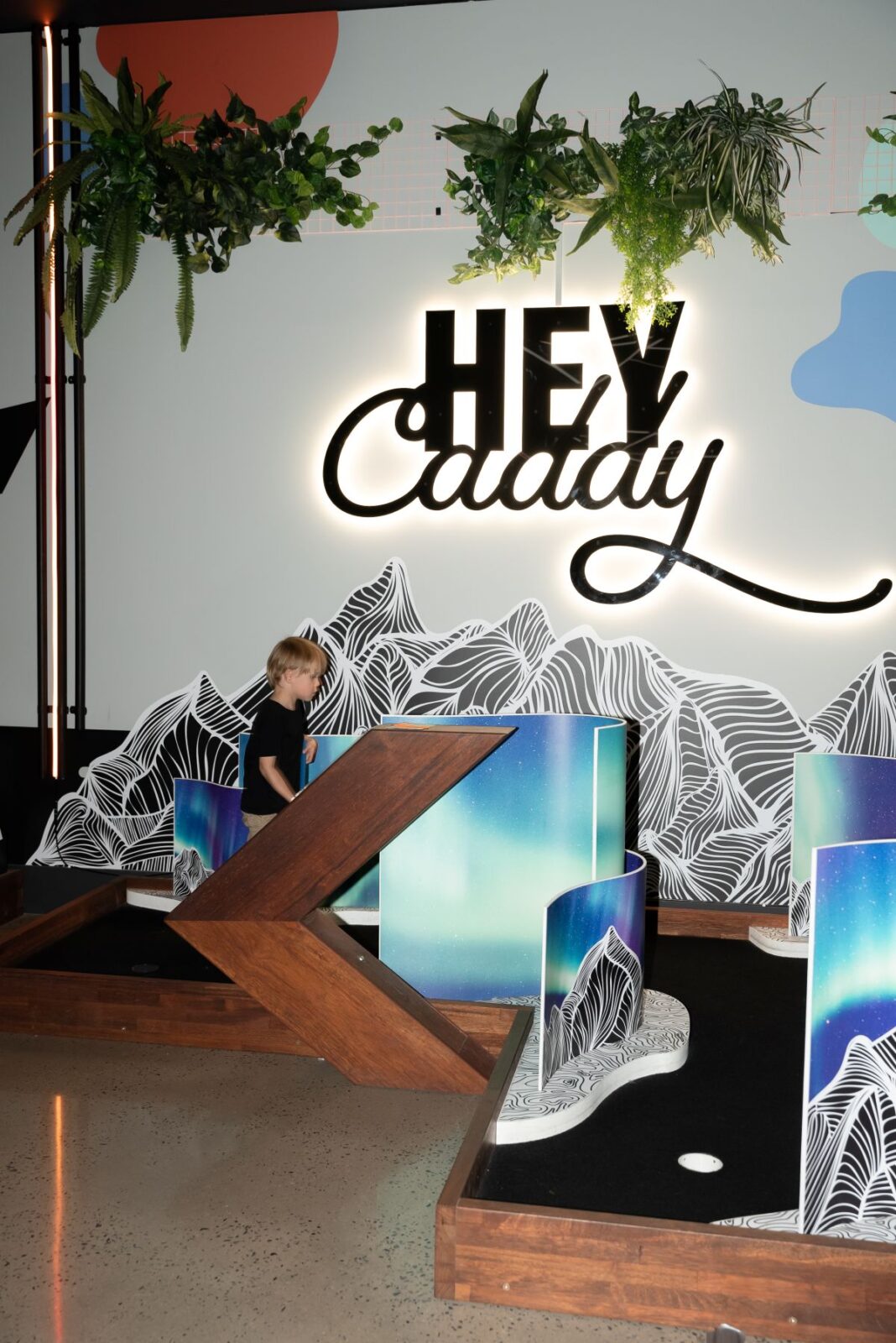 A child joyfully plays mini golf in front of a sign that reads "Hey Caddy," surrounded by colorful course decorations.