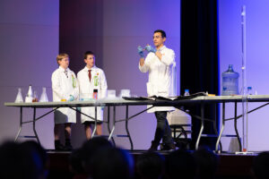 A group of people gathers around a table, with a man in a lab coat leading the discussion.