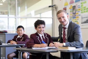 A man in a suit and tie sits at a desk with two boys, engaged in conversation and sharing ideas.